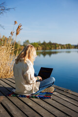 Woman working on laptop while sitting on wooden pier near water, enjoying freedom of remote job....