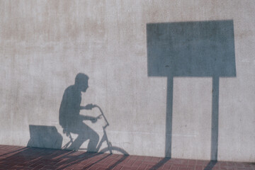 Shadow of delivery man on a bike passing through shadow of rectangular street sign cast on a concrete wall. Conceptual abstract background of the daily grind. Everyday cycle of making a living.
