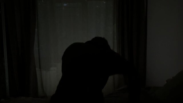 Angry man sitting on a bed in a dark room aggressively hitting a pillow in frustration.