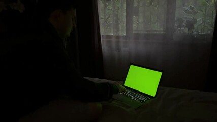 Man sitting on bed typing on laptop with green screen for customizable content.
