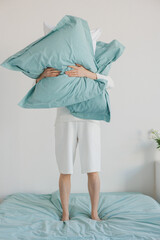 A person is currently standing on a bed while holding a soft pillow directly in front of their face, creating a playful atmosphere