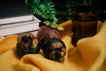 Lush still life set up with three purple Mangosteen, a stack of books, green vines, vibrant yellow felt cloth, glass with water. Close-up of sweet and tangy tropical fruit native to Southeast Asia.