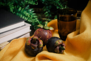 Lush still life set up with three purple Mangosteen, a stack of books, green vines, vibrant yellow felt cloth, glass with water. Close-up of sweet and tangy tropical fruit native to Southeast Asia.