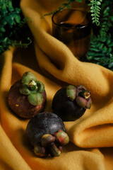 Dramatic and lush still life set up with three purple Mangosteen, green vines, vibrant yellow felt cloth, and a glass with water. Close-up of sweet and tangy tropical fruit native to Southeast Asia.