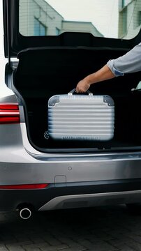 Close-up of a hand carefully placing a silver, hard-shell suitcase into the open trunk of a gray car, suitable for travel vlogs or car review visuals