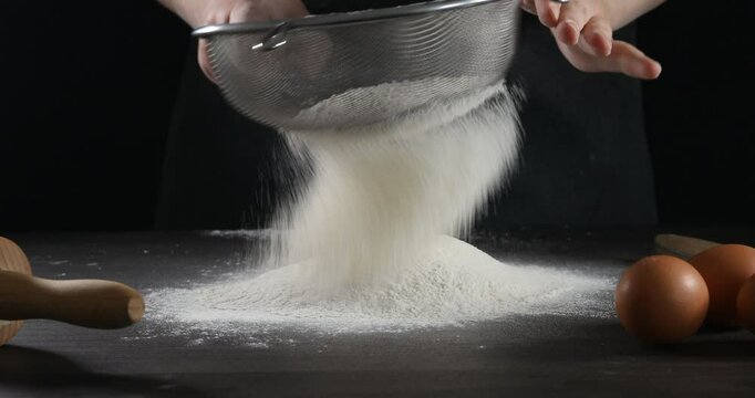 Woman sieving flour, making well and breaking egg into it at dark wooden table against black background, closeup
