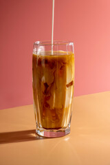 Milk pouring into ice coffee on pink background.