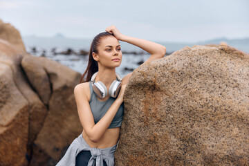 Young woman with headphones leaning against a rock by the sea, portraying relaxation and enjoyment of nature in activewear