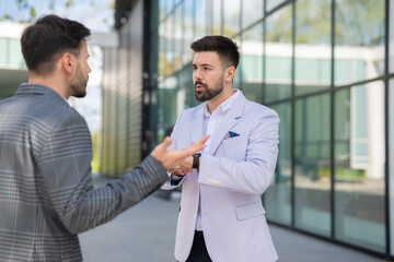 Two businessmen having a serious discussion outdoors, one checking the time while the other gestures expressively.
