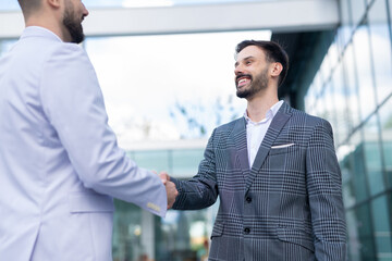 Two businessmen shaking hands outdoors in front of a modern office building, smiling and resolving a conflict or finalizing a business agreement.

