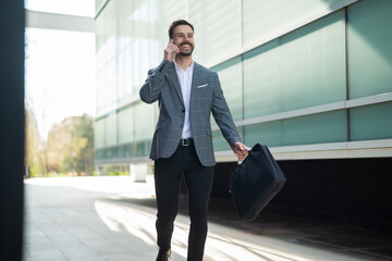 Cheerful businessman walking outside a modern office building, holding a briefcase and talking on a mobile phone with a big smile.