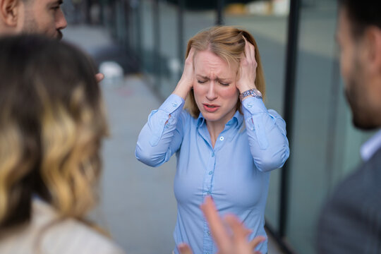 Young professional woman in blue shirt holding her head and covering her ears while coworkers argue around her. Concept of mental pressure, workplace harassment, stress and team conflict.