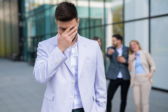 Sad businessman feeling embarrassed while colleagues laugh behind him