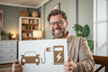 Fototapeta premium Smiling man showing electric car charging station symbol at home