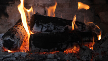 Warm and inviting close-up of burning logs in a fireplace. Bright orange and yellow flames dance around the charred wood, illuminating the surrounding hearth with a soft glow.