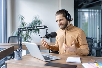 A man with a beard uses a microphone and laptop, engaging in a live podcast recording in an office...