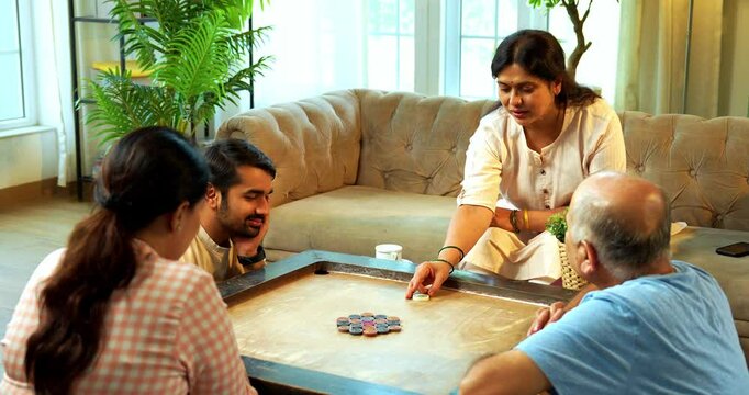 Playing carom or carrom tabletop game together, Indian family of four with senior parents and young adult son and daughter enjoying fun bonding time in cozy living room at home indoor leisure activity