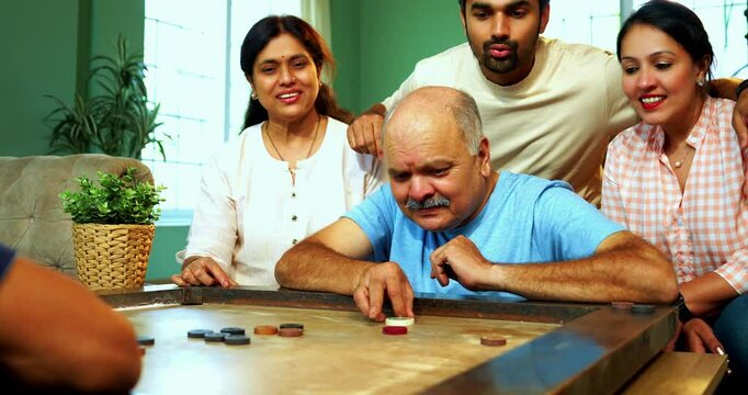 Playing carom or carrom tabletop game together, Indian family of four with senior parents and young adult son and daughter enjoying fun bonding time in cozy living room at home indoor leisure activity