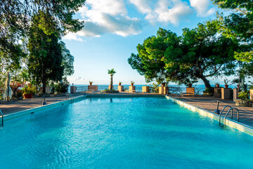 swimming pool with blue water in a hoter resort park during summer tourism season with green trees and blue sea water and cloudy sky on background