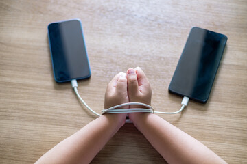 Child's Hands tied with charging cables between two smartphones on wooden table, symbolizing smartphone addiction and dependency in modern digital life. Conceptual image for technology overuse