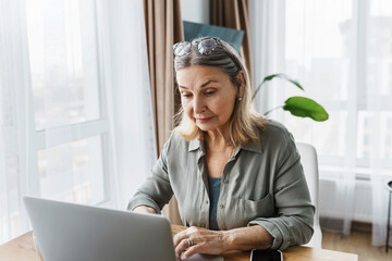 Indoor image of aged businesswoman working on her day-off sitting at table using laptop, solving current issues of her small business, looking at screen with serious tired facial expression