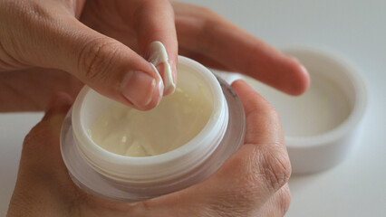 Person applies cream from a small container, demonstrating a skincare routine in natural light at...