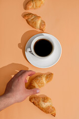 Hand holding fresh croissant on orange background with coffee cup and fresh pastry on plate....