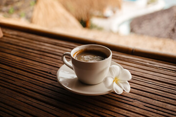 Hot black coffee in a white mug with frangipani flower. Black coffee on a bamboo table. Cup of strong coffee in the morning on vacation	