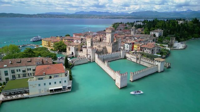 Drone flyaway from Scaliger Castle in Sirmione, Italy, revealing turquoise waters of Lake Garda, colorful houses, and historic walls on a sunny spring day.
