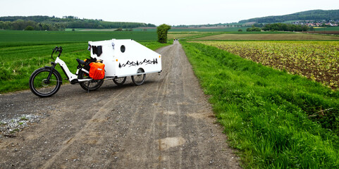 Electric bicycle pulling micro caravan on gravel road through countryside fields