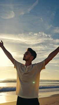 A joyful oung man raises both arms toward bright sunrise over the sea in a vertical video. concept of gratitude, hope, and spiritual awakening.