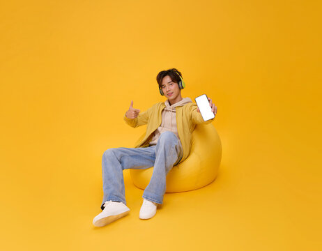 Happy young asian man listening to music with headphone and showing mobile phone sitting bean bag isolated on yellow background. People lifestyle concept.