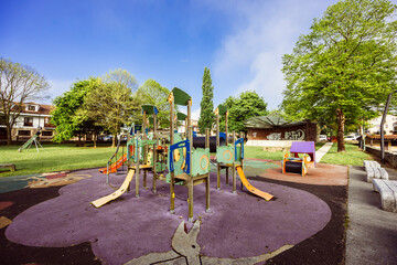 Outdoor playground featuring colorful slides, climbing frames, and soft rubber flooring, surrounded by green trees and a clear blue sky