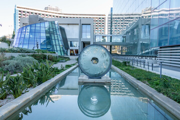 A modern architectural scene featuring a glass building with a reflective pool and a circular...
