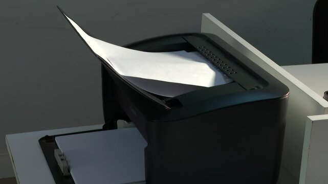 A black office printer sits on a white table, with a blank sheet of paper sticking out. Ready to print important documents for work.