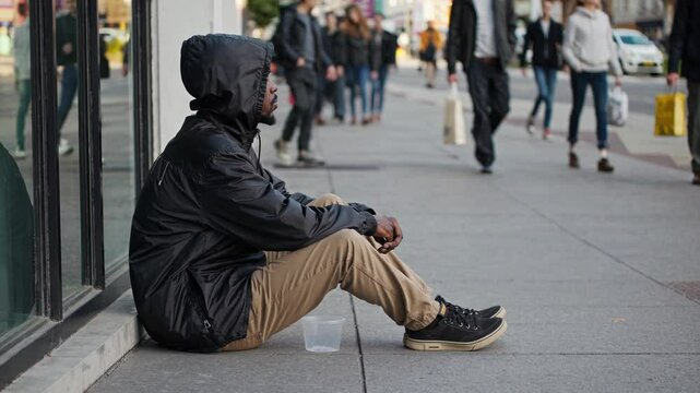 Homeless young male sitting on urban sidewalk, holding donation cup while surrounded by busy city pedestrians, embodying economic hardship and social isolation