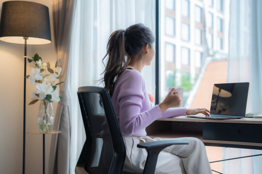 Young Asian businesswoman is working from home, sitting on an ergonomic chair in front of her laptop, drinking coffee and looking out the window of her apartment during a break