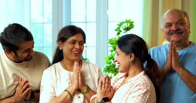 Standing in namaskar pose and smiling at camera, Indian family of four including senior parents and young adult son and daughter greeting with folded hands in cozy modern home interior setting