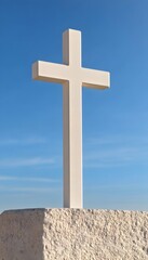 A simple white cross stands tall against a clear blue sky, atop a light-colored stone base