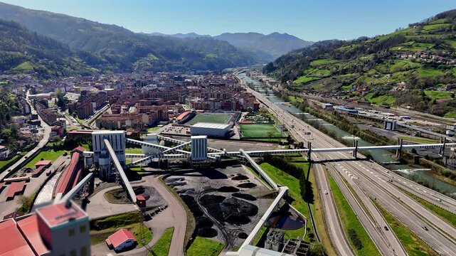 Drone panoramic view of a city nestled in a mountain valley with highways, industrial plant, river, and dense residential zones in Mieres, Asturias.