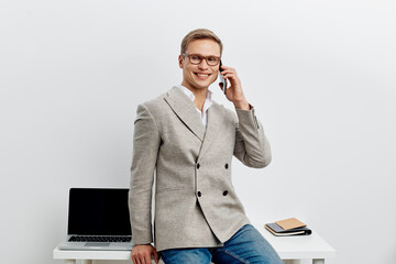smiling young man talking smartphone wear casual blazer glasses sit near laptop desk isolated on...
