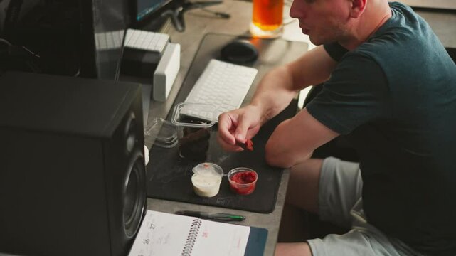 aerial view of remote worker dipping dark bread snack into red sauce while looking at food with cup of beer placed near computer keyboard monitor speaker and open notebook on home office desk