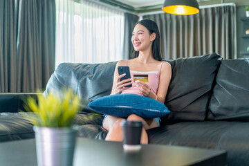 Asian Woman Holding Credit Card and Smartphone on Sofa. Young Lady Shopping Online or Banking Digitally from Home Living Room. © StockPhotoRepublic