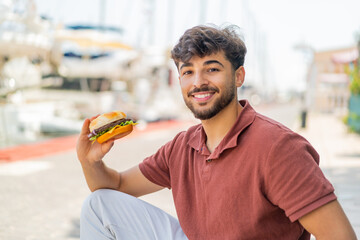 Young Arabian handsome man holding a burger at outdoors smiling a lot