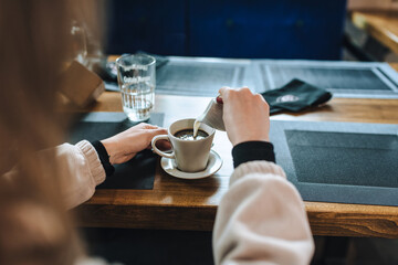Young woman adding milk to coffee in cup on wooden table in cafe. Food photography.