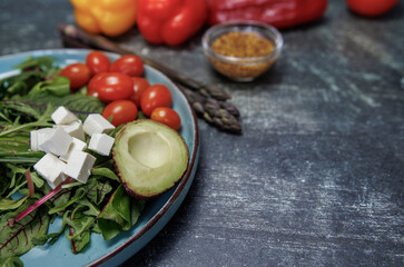 The concept of healthy eating and veganism. Fresh vegetables and herbs on a dark surface. Ingredients include avocado, cherry tomatoes, bell peppers, asparagus, parsley and grain mustard. 