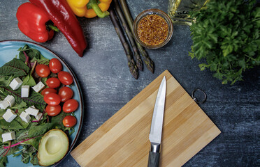 Fresh salad with avocado, cherry tomatoes, cubes of white cheese and herbs on a dark textured surface. Nearby are bell peppers, asparagus, mustard seeds, a wooden board with a knife and a bottle 