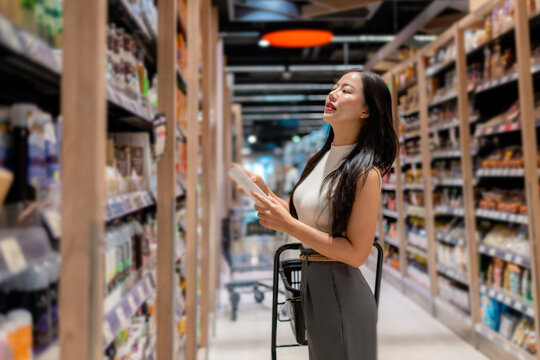 Customer choosing products, reading label on goods in grocery store, holding a cart, walking through the aisle and searching for food