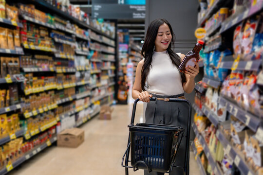 Asian customer reading ingredients of a product pushing a shopping cart in a grocery store - Powered by Adobe