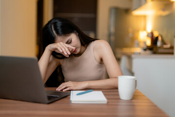 Young Asian woman rubbing her tired eyes while working from home at her laptop at night